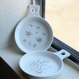 May include: Two white ceramic dishes with handles. Both dishes have a floral design with red and green accents.