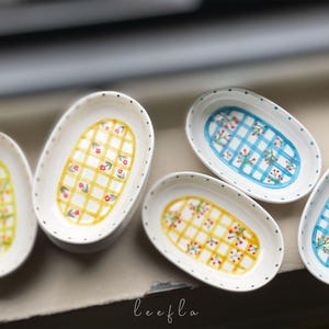 May include: A collection of oval-shaped ceramic dishes. Each dish features a white base with a hand-painted design. The designs include yellow and blue checkered patterns with floral accents. The edges are dotted with small black dots.