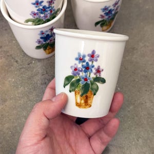 May include: White ceramic cups with a floral design featuring blue and purple flowers in a gold pot. The cups have a simple, rounded shape and are stacked on a gray surface. The cups are decorated with a hand-painted floral design.