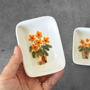 May include: White ceramic dish with a hand-painted floral design. The design features orange flowers in a brown pot with green leaves. The dish is rectangular with rounded corners. Another similar dish is visible.