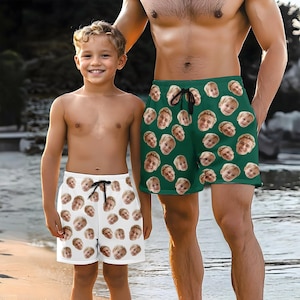 May include: Two people wearing matching swimming trunks with a pattern of faces. The trunks are green and white. The people are standing on a beach.