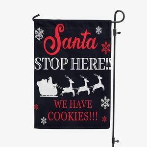May include: A black garden flag with the text "Santa STOP HERE!! WE HAVE COOKIES!!!" in red and white. The flag features a graphic of Santa's sleigh and reindeer, with snowflakes. The flag is displayed on a black metal stand.