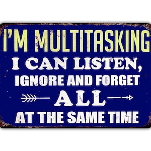 May include: A blue metal sign with white text that reads "I'm multitasking, I can listen, ignore and forget all at the same time". The sign has a distressed look and features two arrows pointing in opposite directions.