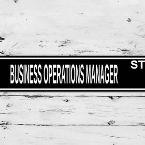 Pode incluir: Placa preta e branca com o texto "BUSINESS OPERATIONS MANAGER ST." em um fundo de madeira branca.
