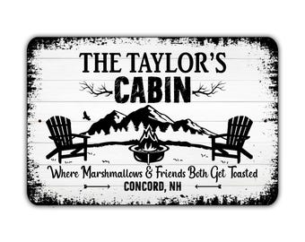 May include: A rectangular, distressed white sign with black text and graphics. The sign reads "THE TAYLOR'S CABIN" above a mountain scene with a campfire and two chairs. Below, it says "Where Marshmallows & Friends Both Get Toasted" and "CONCORD, NH."