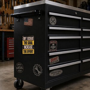 A black workbench with various stickers and labels on it, including a motivational quote and a warning about the importance of the effort.