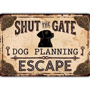 May include: A rectangular metal sign with a distressed cream background featuring a black silhouette of a dog. The text reads SHUT THE GATE DOG PLANNING ESCAPE. The sign has decorative bronze-colored corner brackets with bone motifs.