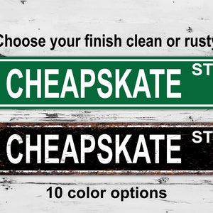 May include: Two metal street signs with the text "CHEAPSKATE ST" in white letters. The top sign is green with a clean finish. The bottom sign is black with a rusty finish. The text "Choose your finish clean or rusty" appears above the signs. The text "10 color options" appears below the signs.