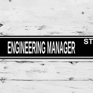 Peut inclure: Un panneau noir et blanc avec le texte "ENGINEERING MANAGER ST." sur un fond en bois blanc.