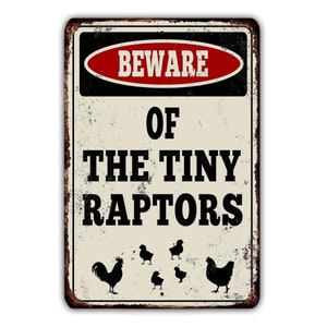 Pode incluir: Um sinal de metal de estilo vintage com um sinal de advertência que diz "Cuidado com os pequenos raptors". O sinal tem uma borda vermelha e preta com texto branco. Há cinco silhuetas pretas de galinhas no sinal.