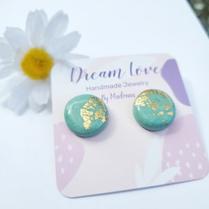 Op de afbeelding: Turquoise oorstekers met goudblad accenten. De ronde oorbellen worden gepresenteerd op een kaart met de tekst "Dream Love Handmade Jewelry by Madonna". Een witte madelief met een gele kern staat op de achtergrond.