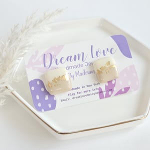 May include: Square cream-colored earrings with gold leaf accents. The earrings are displayed on a white and gold-rimmed hexagonal dish, resting on a card with the text "Dream Love" and other details.