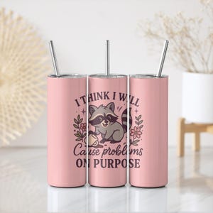May include: Three pink insulated tumblers with silver straws. Each tumbler features a cartoon raccoon illustration with the text "I THINK I WILL Cause problems ON PURPOSE". The design includes floral and star accents, creating a playful aesthetic.