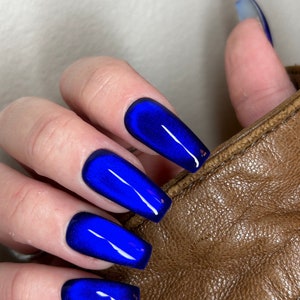 May include: Close-up of a hand with long, blue, cat-eye nails. The nails are a deep, rich blue with a subtle shimmer. The hand is resting on a brown leather surface.