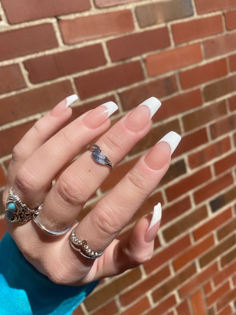 May include: Close-up of a hand with long, white, almond-shaped nails with a French manicure. The nails are painted with a light pink base and a white tip. The hand is wearing a silver ring with a leaf design.
