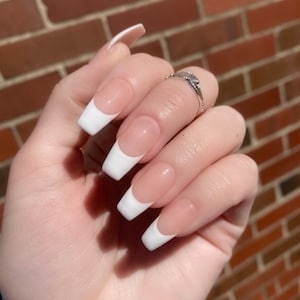 May include: Close-up of a hand with long, white-tipped, almond-shaped nails. The nails are painted with a classic French manicure, featuring a white tip and a nude base.