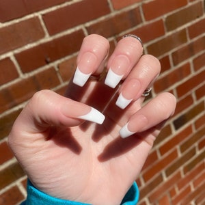 May include: Close-up of a hand with long, white-tipped, almond-shaped nails. The nails are painted with a nude base and a white tip, creating a classic French manicure.