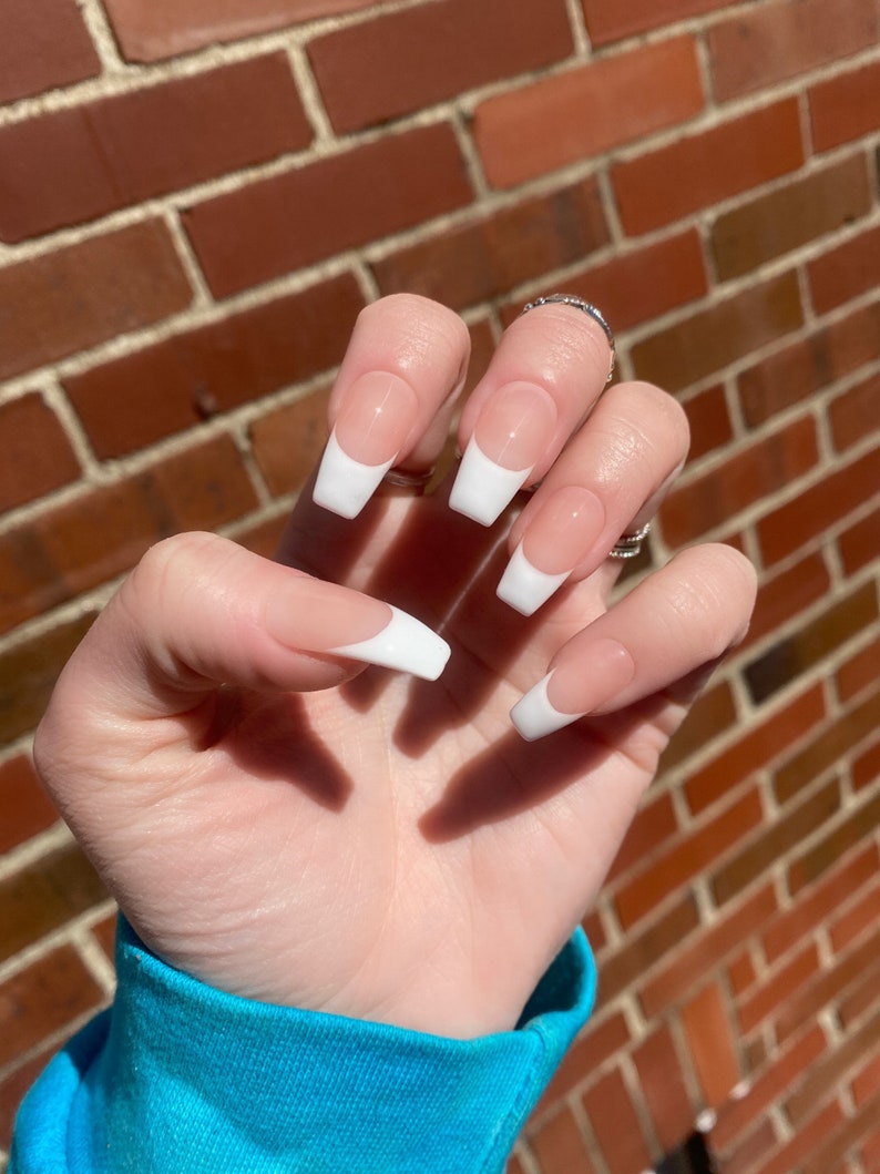 May include: Close-up of a hand with long, white-tipped, almond-shaped nails. The nails are painted with a nude base and a white tip, creating a classic French manicure.
