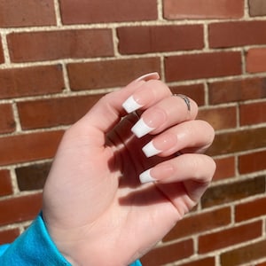May include: Close-up of a hand with white french manicured nails. The nails are long and oval shaped. The hand is against a brick wall background.