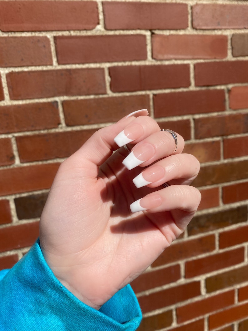 May include: Close-up of a hand with white french manicured nails. The nails are long and oval shaped. The hand is against a brick wall background.