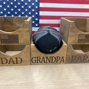 May include: Wooden storage boxes with engraved text reading "DAD", "GRANDPA", and "PAPA". Each box has a cutout design and an engraved American flag. A black and gray baseball cap rests inside one of the boxes. The background features a wooden American flag.