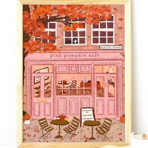 May include: A framed print of the Pink Pumpkin Café, a pink building with a cafe sign and outdoor seating. The artwork features a fall scene with orange leaves and pumpkins. The cafe's name is displayed above the entrance.