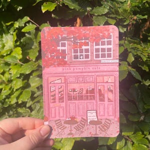 Puede incluir: Ilustración rosa de un café con el texto "pink pumpkin café" y "HARVEST STREET". El café tiene mesas y sillas en el exterior. La imagen se sostiene frente a un fondo de hojas verdes.