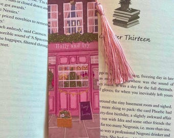 May include: A pink bookmark with a festive illustration of a shop called "Holly and Ivy". The shop has a pink facade with a window, a door, and a sign that says "Merry Christmas".
