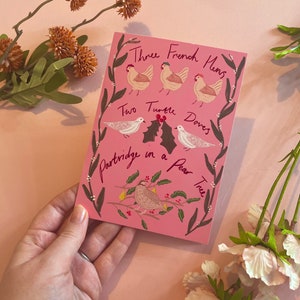May include: A pink greeting card with illustrations of chickens, doves, and a partridge on a pear tree. The card features the text "Three French Hens," "Two Turtle Doves," and "Partridge in a Pear Tree" in a decorative font.