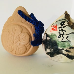 May include: Two small ceramic ornaments, one brown with a raised floral design and a blue cord, the other white with black and green brushstrokes and Chinese characters.