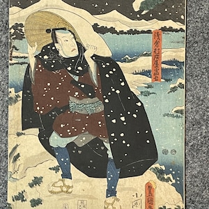 Puede incluir: Una impresión en madera que representa a un hombre con una túnica negra y un sombrero de paja caminando por un paisaje nevado. La impresión está titulada "陳倉村慶蛋" en caracteres japoneses.