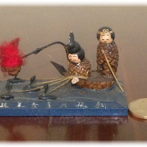 May include: A wooden figurine of two people in traditional Japanese clothing, sitting in a boat made of pine cones. The boat is on a blue wooden base with Japanese characters.