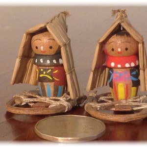 May include: Two wooden dolls with painted faces and colorful clothing are wearing straw hats. The dolls are standing on wooden bases.