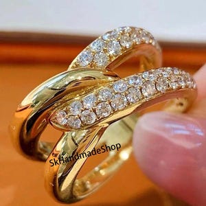 May include: A gold ring with a double band design, featuring a row of small clear stones around the edge of each band. The ring is a modern and elegant piece of jewelry.