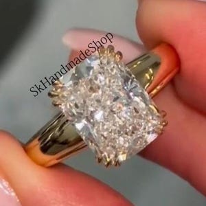 May include: A gold ring with a large, rectangular, clear gemstone. The ring is held between two fingers. The text "SkHandmadeShop" is visible on the ring.