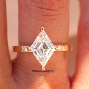 May include: A gold ring with a clear, kite-shaped gemstone as the focal point. The band is plain gold, with small, clear accent stones either side of the main stone. The ring is displayed on a person's finger.