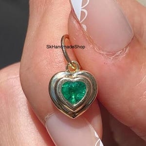 May include: A gold heart-shaped pendant featuring a central green gemstone. The pendant has a smooth, polished gold frame surrounding the heart-shaped green stone. The text SkHandmadeShop is visible on the small gold jump ring at the top.