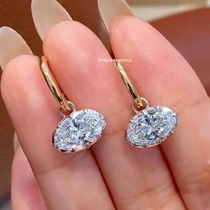 Op de afbeelding: Een paar gouden oorringen met heldere, ovaal geslepen stenen. Elke oorbel heeft een gepolijste gouden ring verbonden met een in klauwen gezette ovale steen die het licht reflecteert. De tekst SkHandmadeShop is zichtbaar in het midden tussen de oorbellen.