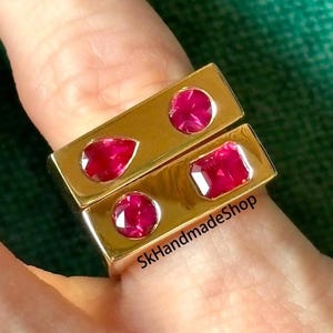 May include: Two gold-colored rings with rectangular bands, each featuring vibrant red gemstones. One ring has three round and one pear-shaped stone, while the other has two round and one square-cut stone. The rings are on a finger. The text "SkHandmadeShop" is visible.