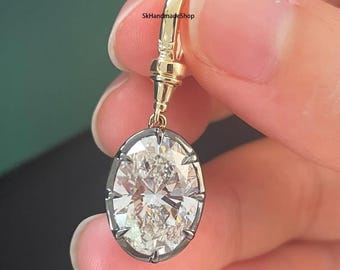 May include: An oval-shaped diamond pendant earring with a gold-coloured top and a dark metal frame. The diamond has a brilliant cut, reflecting light. The earring is held up by a gold-coloured hook. The text "SkHandmadeShop" is visible.