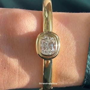 Puede incluir: Un brazalete dorado con un gran diamante de talla cuadrada en el centro. El diamante está engastado en un bisel. El brazalete es de diseño clásico, adecuado para el uso diario o para ocasiones especiales.