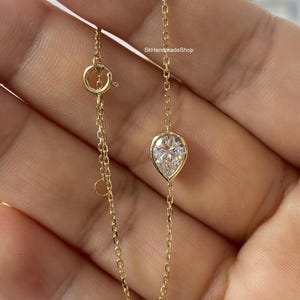 May include: A delicate gold chain necklace with a pear-shaped diamond pendant. The pendant is set in a gold bezel and sparkles. The necklace has a lobster clasp closure. The text "SkHandmadeShop" is visible.
