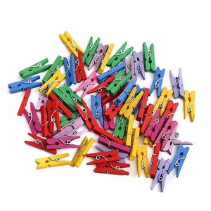 Pode incluir: Uma coleção de pequenos prendedores de roupa de madeira em várias cores, incluindo vermelho, amarelo, verde, azul, rosa e roxo. Os prendedores estão espalhados sobre uma superfície branca, mostrando suas cores vibrantes e design simples.