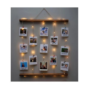 Puede incluir: Un expositor de fotos de madera con luces de cadena y pinzas para sujetar fotos. Las fotos son de una familia y tienen fechas escritas en ellas. El expositor está colgado en una pared.