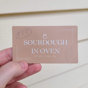 以下が含まれることがあります： 「SOURDOUGH IN OVEN DO NOT TURN ON」と白文字で書かれた茶色のカード。カードにはパンと瓶のイラストが描かれている。