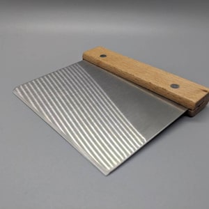 May include: A small, rectangular metal pastry scraper with a wooden handle. The handle has two small metal rivets. The metal blade has a textured surface.