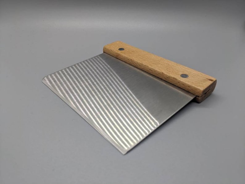 May include: A small, rectangular metal pastry scraper with a wooden handle. The handle has two small metal rivets. The metal blade has a textured surface.