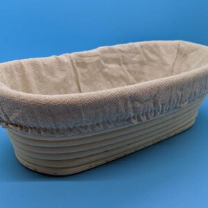 May include: A light brown oval-shaped bread proofing basket with a removable linen liner. The basket is made of woven rattan.