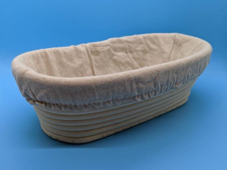 May include: A light brown oval-shaped bread proofing basket with a removable linen liner. The basket is made of woven rattan.
