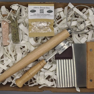 Puede incluir: Una caja blanca llena de papel de embalaje marr&oacute;n y una variedad de herramientas para hornear. Las herramientas incluyen un rodillo de madera, un raspador de masa, un batidor de masa, dos peque&ntilde;os frascos de vidrio y una peque&ntilde;a bolsa de papel marr&oacute;n con un c&oacute;digo QR. La bolsa tiene el texto "John's Bread Box" y una direcci&oacute;n de sitio web.